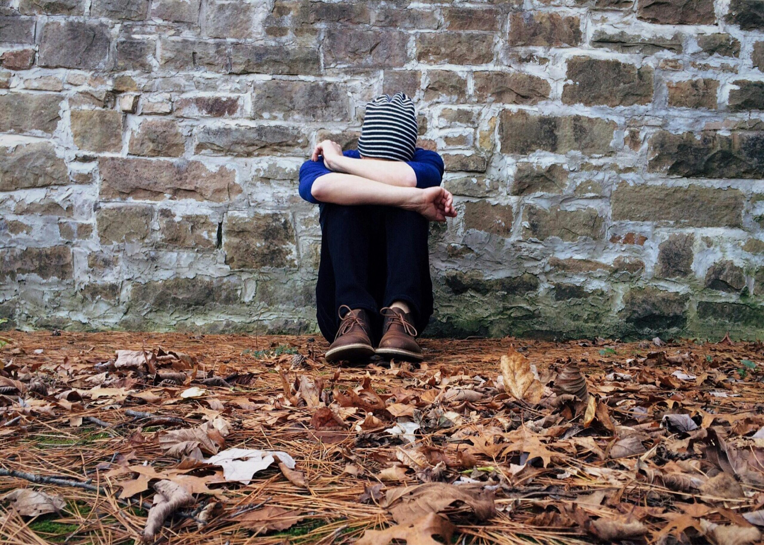 A solitary figure sits against a stone wall surrounded by fallen leaves, expressing isolation.