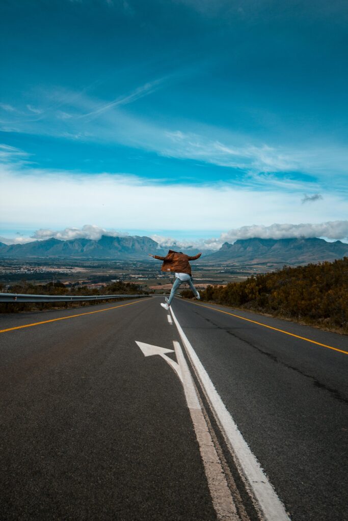 A person jumps on a remote mountain highway under a vibrant blue sky, conveying freedom and adventure.