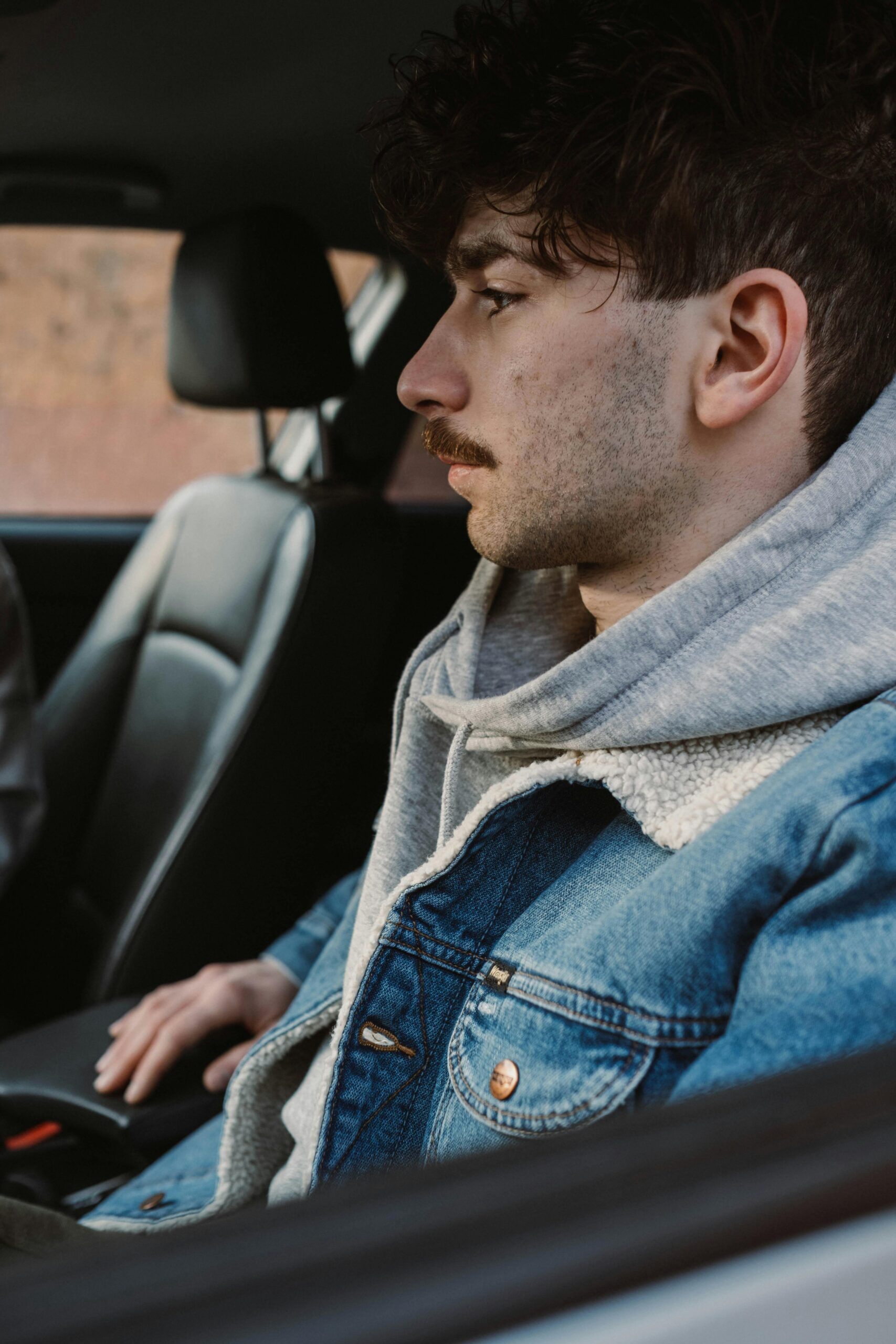 Young man with denim jacket and mustache sitting thoughtfully in a car interior.