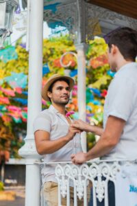 A gay couple holding hands outdoors, showcasing love and romance amid colorful decorations.